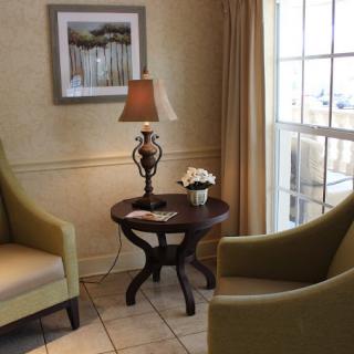 A cozy living room corner with two armchairs, a round wooden table, and a lamp beneath a framed picture by the window.