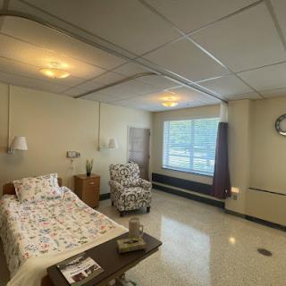 A well-lit and comfortable hospital patient room featuring a bed, armchair, and large window.