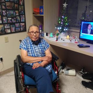 An elderly person sitting in a wheelchair, surrounded by holiday decorations and watching TV.