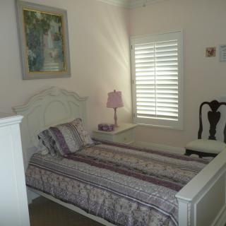 A cozy bedroom with a single bed, lamp, painting, and window shutters.
