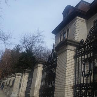 An elegant brick building with an ornate wrought iron gate, framed by bare trees under a cloudy sky.