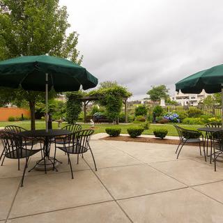 An outdoor patio area with tables, chairs, and green umbrellas in a lush garden setting on a cloudy day.