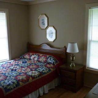 A cozy bedroom with a colorful quilt on the bed, a nightstand with a lamp, and two windows providing natural light.