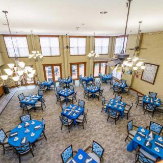 A spacious dining area with multiple tables covered in blue tablecloths, surrounded by chairs and well-lit by chandeliers.