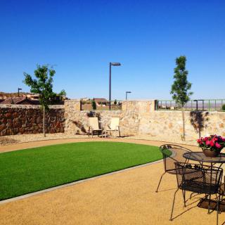 A well-maintained backyard with artificial grass, stone walls, and patio furniture under a clear blue sky.