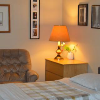 A cozy bedroom with a soft armchair, lit lamp on the nightstand, and framed pictures on the wall.