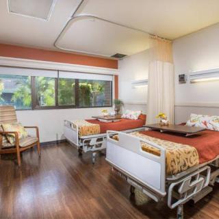 A well-lit hospital room featuring two patient beds, a large window, and comfortable furniture.