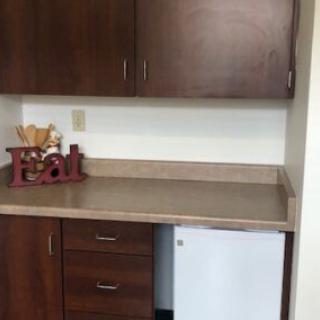 A small kitchenette with wooden cabinets and a countertop, featuring a mini fridge and decorative plants.
