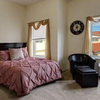 A cozy bedroom featuring a neatly made bed, a comfortable chair, and two windows with decorative curtains.