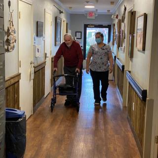 An elderly person using a walker is accompanied by a caregiver in a hallway of an assisted living facility.