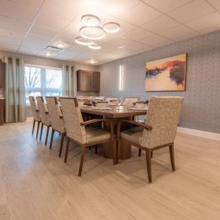 A modern dining room featuring a large wooden conference table, surrounded by upholstered chairs, with stylish chandelier lighting and artwork on the wall.