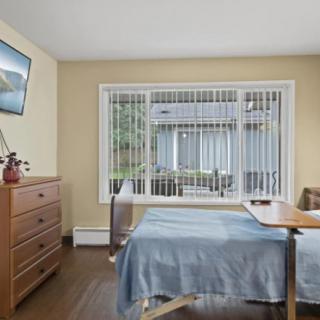 A cozy bedroom featuring a single bed with a blue cover, a dresser, and a mounted television. The room is well-lit by natural light coming through the large window.