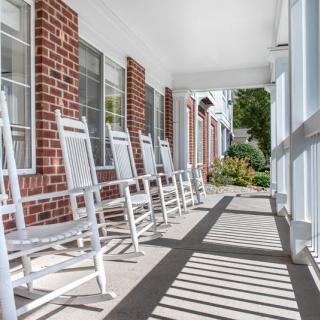 A sunny porch lined with white rocking chairs against a brick wall.
