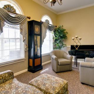 A cozy living room featuring a grandfather clock, comfortable armchairs, and a piano near large windows with elegant drapery.