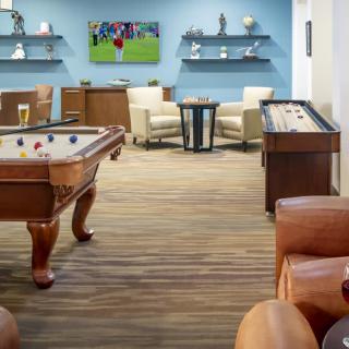 A modern game room featuring a pool table, shuffleboard, comfortable seating, and a television displaying a sports event.