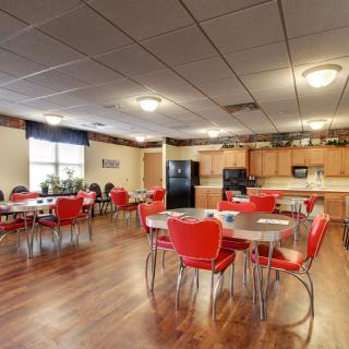 A spacious community dining area with red chairs and wooden tables, featuring a kitchen setup with cabinets and appliances.
