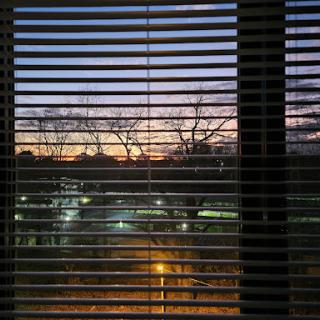 A serene view of the sunset through window blinds, showcasing the transition from day to night in an urban setting.