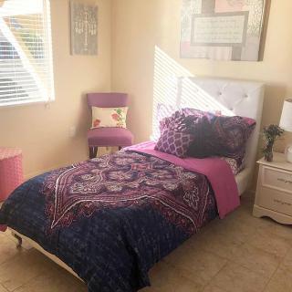 A cozy bedroom with a single bed adorned with purple bedding, a chair in the corner, and sunlight streaming in through the window.