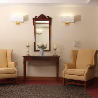 A traditional living room setup with two upholstered chairs, a wooden table, and a mirror above, flanked by wall lamps.