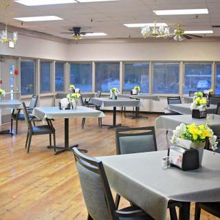 A bright dining area with several tables and chairs, featuring floral centerpieces by large windows.