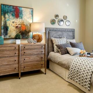 A modern bedroom with elegant furnishings, featuring a textured gray headboard, wooden dresser, abstract artwork, and a cozy lamp.