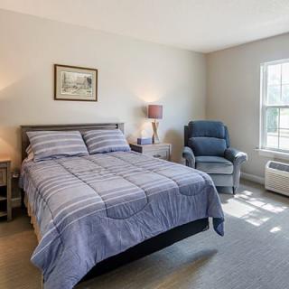 A cozy bedroom with a neatly made bed, a lamp on a nightstand, and a comfortable chair by the window.