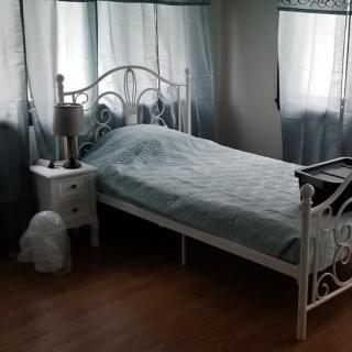 A cozy bedroom featuring a single bed with wrought iron frame, a nightstand with a lamp, and soft curtains lining the windows.