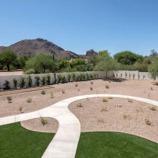 A landscaped desert backyard featuring a winding concrete pathway, sparse vegetation, and distant mountains under a clear blue sky.