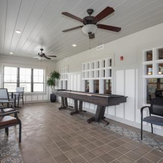 A spacious recreation room featuring a shuffleboard table, ceiling fans, and ample natural light through large windows.