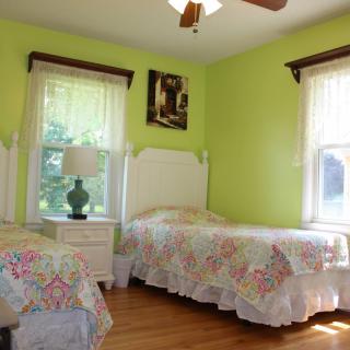 A cozy bedroom with twin beds and green walls, illuminated by natural light from two windows.
