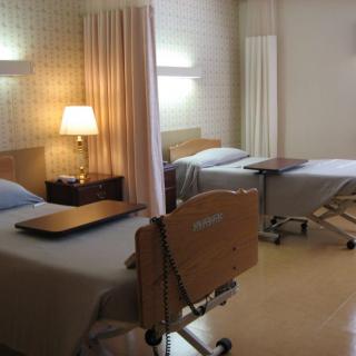 A hospital room with two beds separated by a curtain, featuring bedside tables and a lamp.