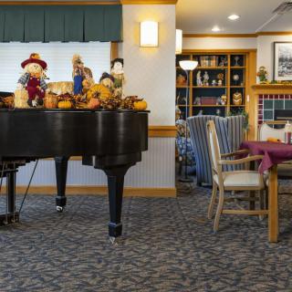 A cozy living room with a grand piano decorated with scarecrows and pumpkins, surrounded by comfortable seating and bookshelves.