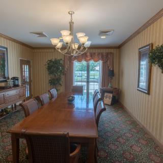A dining room with a long table and chairs, illuminated by a chandelier, featuring large windows with curtains and decorated with artwork and plants.