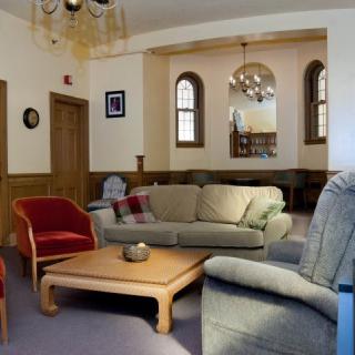 A cozy living room with sofas, chairs, and a coffee table, featuring a chandelier and arched windows in the background.