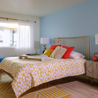 A brightly decorated bedroom with a large bed, colorful pillows, and natural light streaming through the window.