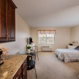 A cozy bedroom interior featuring a bed, table with chairs, cabinetry, and a window providing natural light.