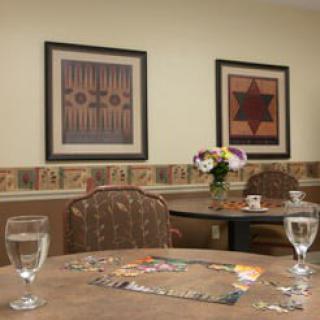 A cozy dining area with tables and chairs, decorated with wall art and flowers, featuring a puzzle in progress.