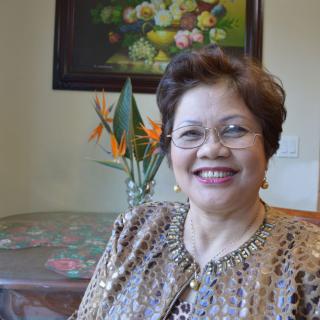 A woman with glasses smiling indoors, sitting near a table with flowers and a painting on the wall.