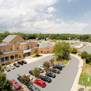 An aerial view of a suburban area with a building and parking lot surrounded by greenery under a cloudy sky.