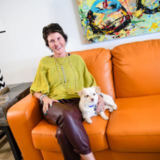 A woman sitting on an orange couch with her small dog, surrounded by colorful artwork.