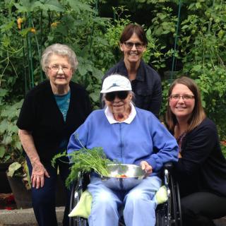 A group of people smiling outdoors in a garden, with one person in a wheelchair holding plants.