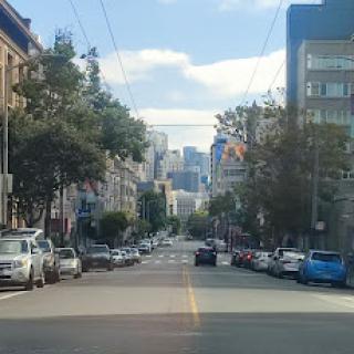 A view of a city street lined with parked cars and buildings under a clear sky.