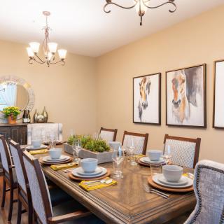 A neatly arranged dining room featuring a wooden table set with plates and cups, accented by artwork on the walls and illuminated by chandeliers.