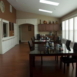 A spacious dining room with wooden flooring, featuring a large table surrounded by chairs, and a kitchen area in the background.