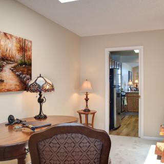 A cozy home office featuring a desk, lamps, and artwork on the wall with a view into the kitchen.