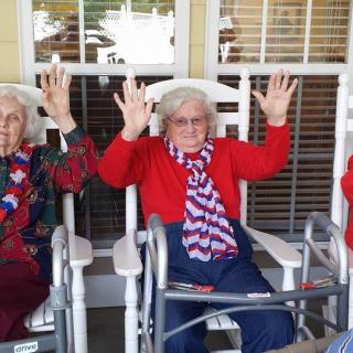 Elderly women in patriotic attire sitting on rocking chairs, raising their hands joyfully.
