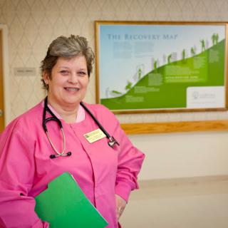A cheerful nurse in pink scrubs holds a green folder in a hospital setting.