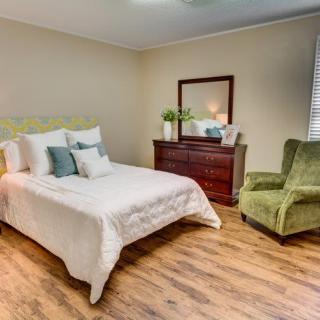 A cozy bedroom with a comfortable bed, green armchair, dresser, and wooden flooring.