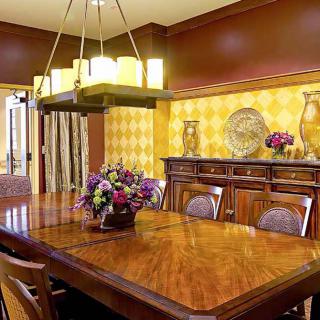 An elegant dining room featuring a wooden table, a modern chandelier, and decorative wallpaper adorned with floral arrangements and decorative plates.
