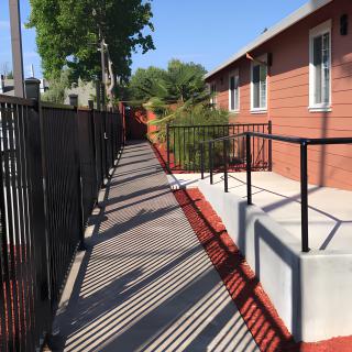 An outdoor ramp alongside a building with a black metal fence and shadows cast on the pathway.